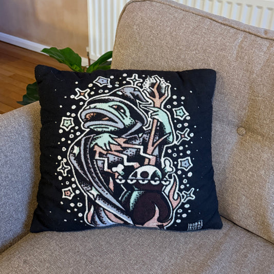 A square black throw pillow featuring a detailed graphic print of a hooded figure with a skull and crossbones motif, resting on a beige fabric armchair in a home setting.